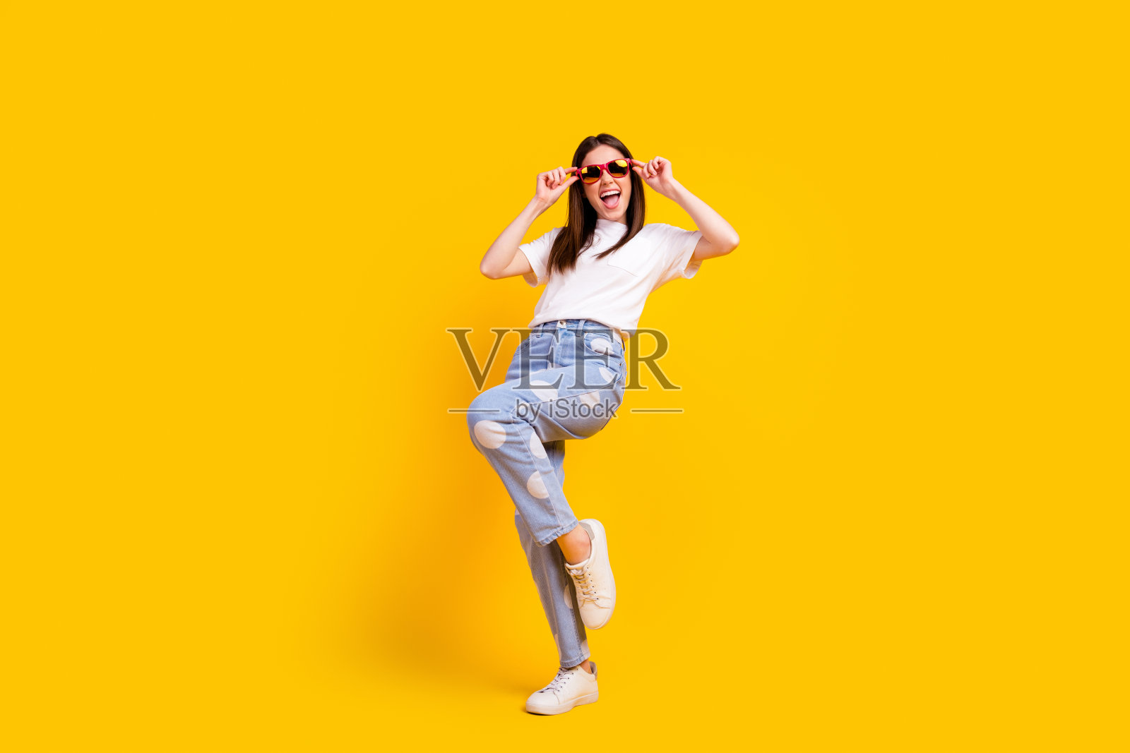 全身照，年轻兴奋的女孩，快乐积极的微笑，嬉皮风格，双手触摸太阳镜，背景为黄色。照片摄影图片