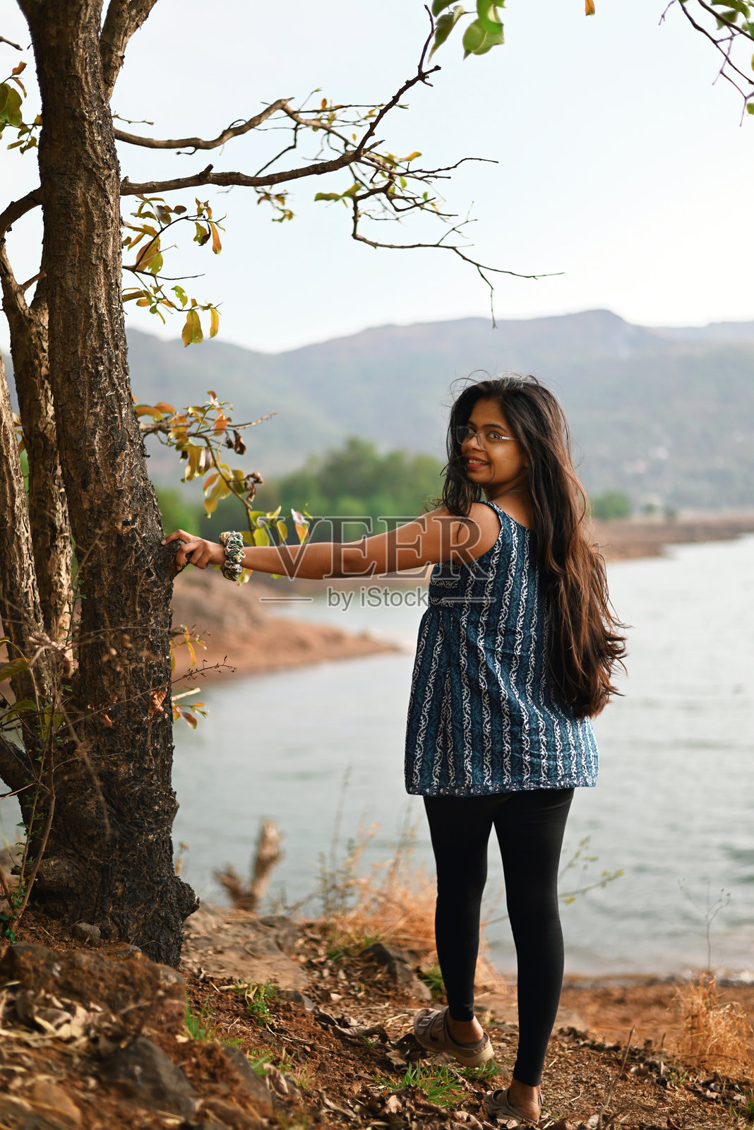 年轻女子在湖边的悬垂树枝下，长发飘逸地站着照片摄影图片