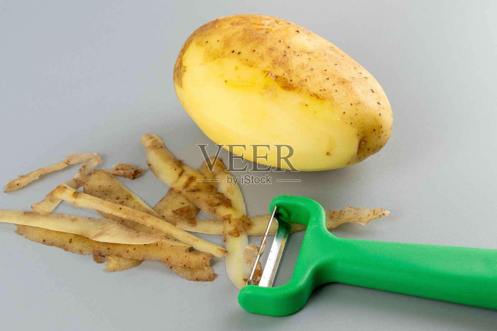 新剥皮的土豆与绿色削皮器在灰色背景上照片摄影图片
