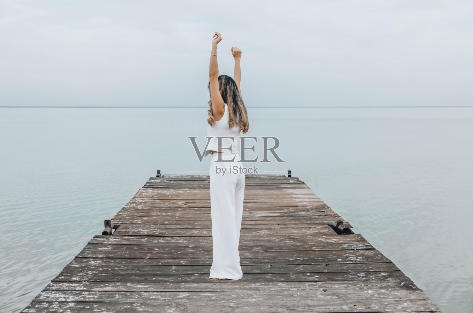 一个无法辨认的女性赤脚站在木栈道上，双手举起，面朝平静的海平面，穿着白色服装，沐浴在淡淡的云层下，展现出正念与健康的氛围，适合旅行博客。照片摄影图片