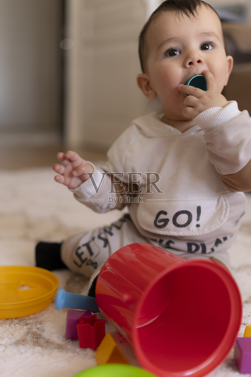 婴儿男孩坐在地毯上，嘴里含着小玩具零件，周围有五彩缤纷的物品，营造出温馨的家居环境，强调早期口腔探索和潜在的窒息危险，旨在提高儿童安全和家长的意识。照片摄影图片