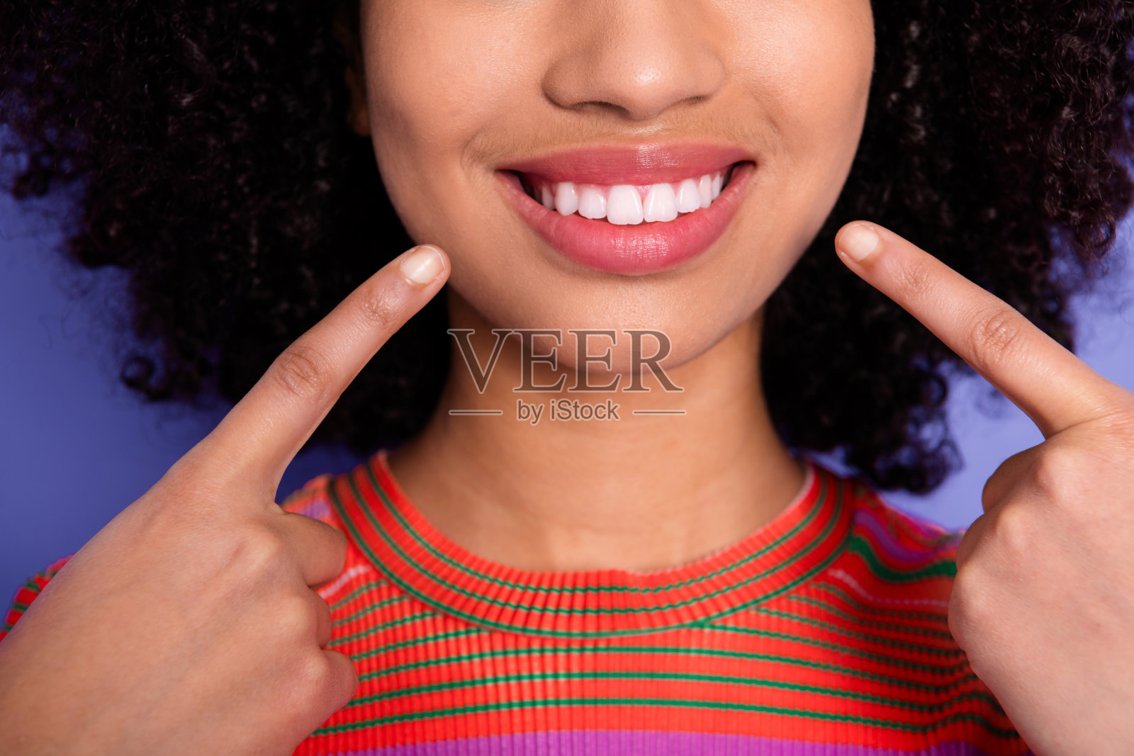 微笑的年轻女性用手指比划，展示她完美的牙齿，背景为鲜艳的紫色。照片摄影图片