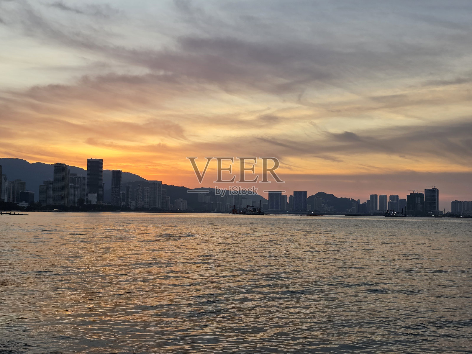 槟城城市天际线在平静的海面上日落时分，天空中的温暖橙色和紫色映照在水面上。照片摄影图片