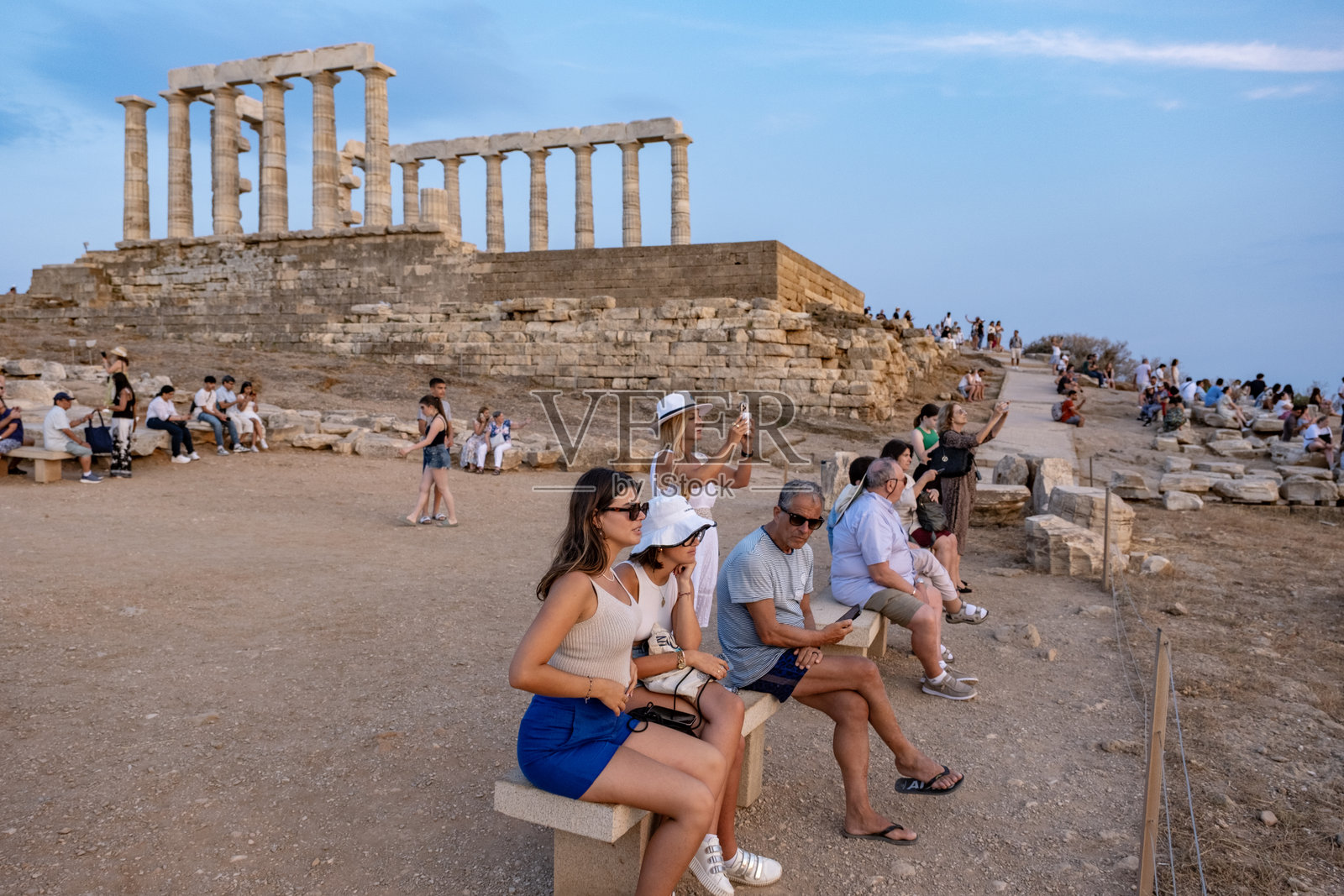 一群游客在希腊苏尼翁角的波塞冬神庙前的长椅上坐着，享受日落时分。照片摄影图片