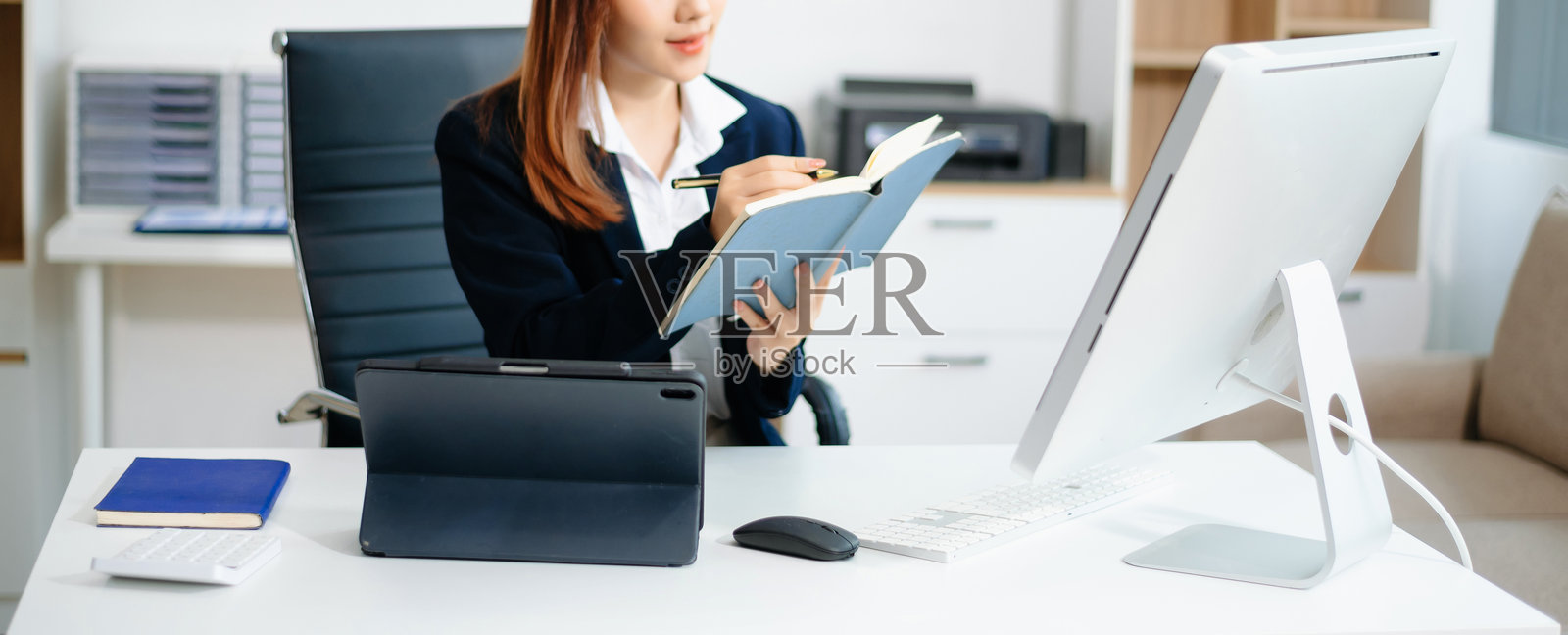 穿着商务套装的亚洲女性办公室工作人员微笑着看向镜头，手边有工作笔记本、平板电脑和笔记本电脑文件。照片摄影图片