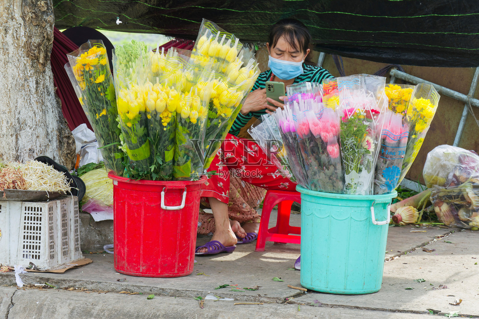 越南街头小贩戴着口罩，一边使用手机，一边在Hoa Phu工业园区出售五彩缤纷的新鲜花卉和农产品。照片摄影图片
