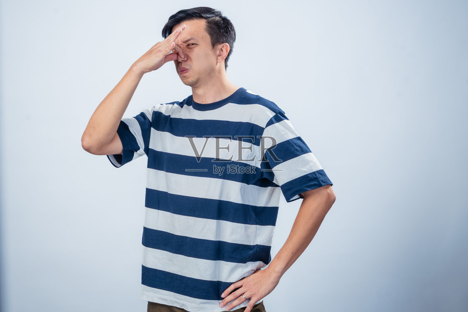 穿着海军蓝和白色条纹T恤的亚洲男子捂住鼻子，吐出舌头，面露厌恶表情，站在简单的浅色背景前，象征着难闻的气味或反应。照片摄影图片