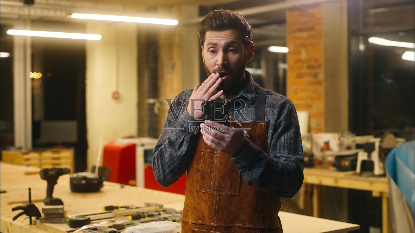 惊讶的白人木工修理工男士在工作室里用手机惊讶地说“哇”，表达了他的情感，庆祝着作为工匠的成功与兴奋。照片摄影图片