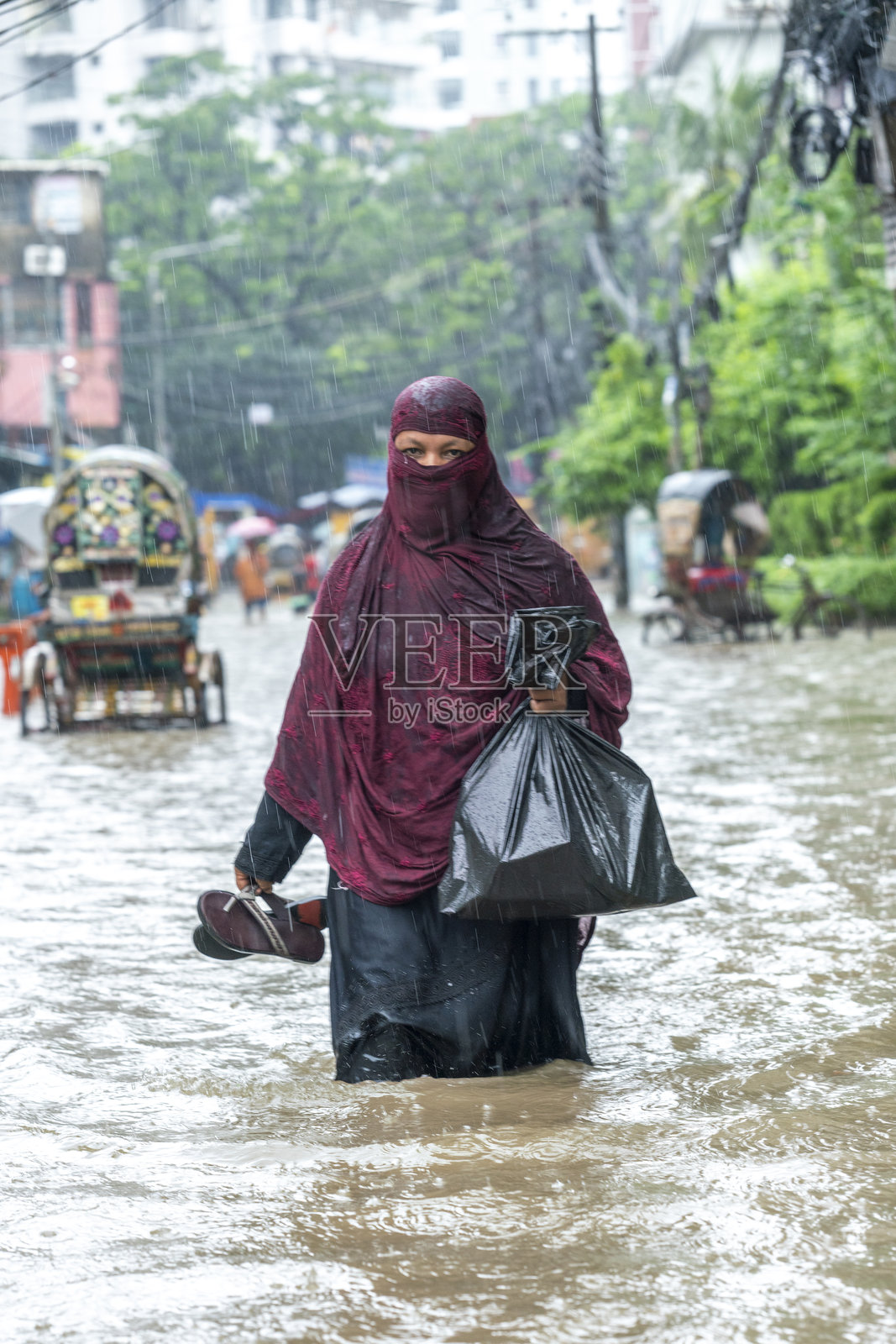 一名女性在城市街道的洪水中涉水,手中提着鞋子和包。水涝是孟加拉国的一个严重问题。照片摄影图片