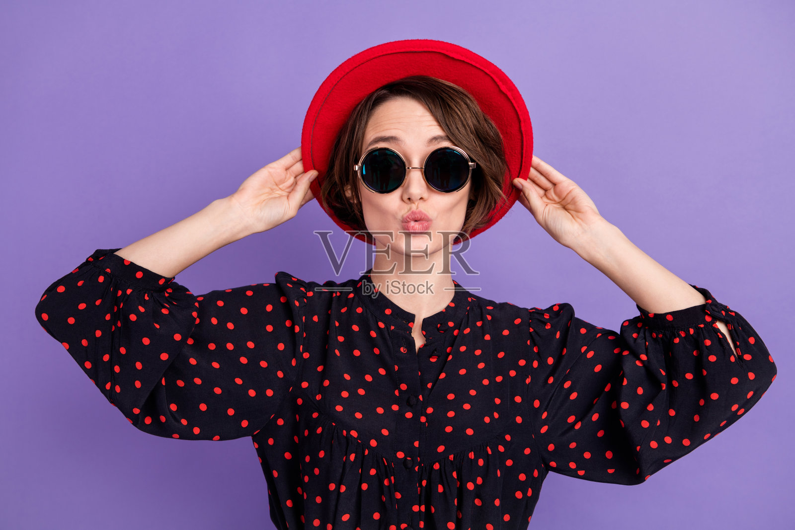 迷人的女孩在明亮的紫色背景下送出空气吻，轻触帽子。照片摄影图片