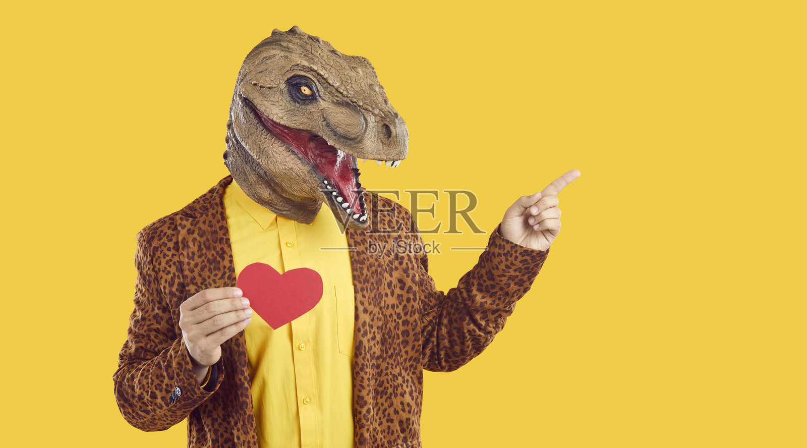 戴着恐龙面具的搞笑男子手持红色的情人节卡片，指向黄色背景上的空白处照片摄影图片