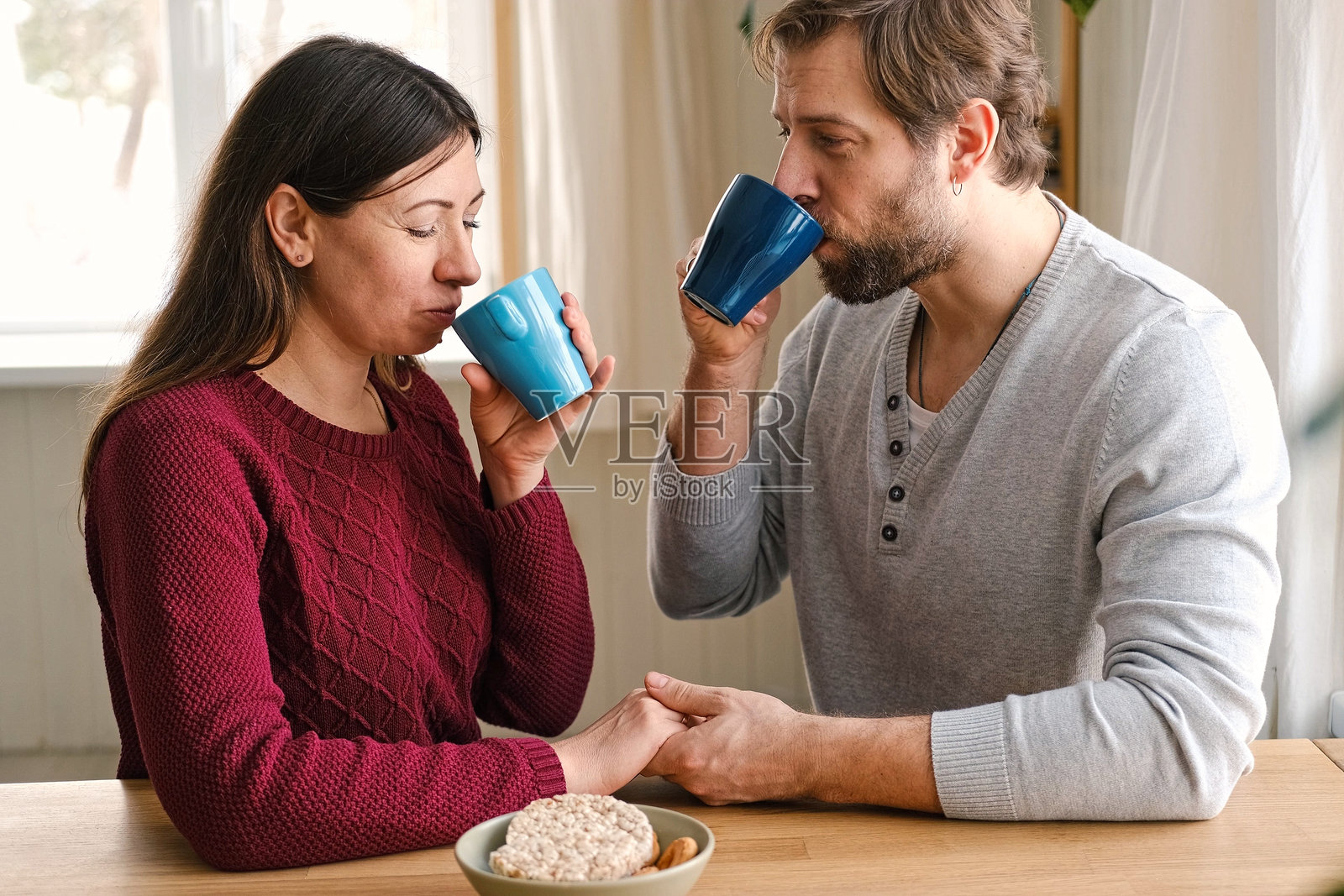温馨的成熟夫妻在家中的互动。丈夫和妻子拥抱，牵着手，一起喝早晨咖啡。男女讨论周末的计划。自然光线。亲密的时刻。照片摄影图片