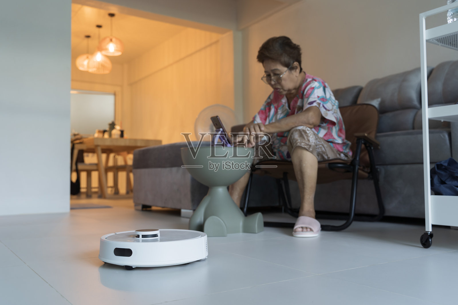 一位年长的亚洲女性在现代客厅中使用智能手机，同时机器人吸尘器在地板上工作。照片摄影图片