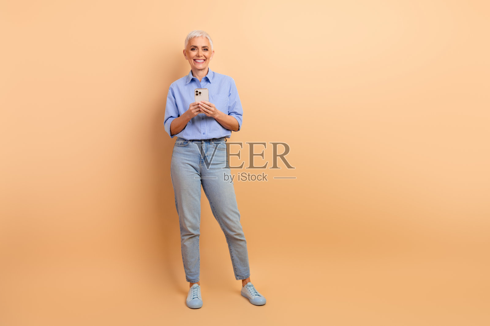 时尚的成熟的女性在米色背景前手持智能手机，穿着蓝色条纹衬衫和牛仔裤，面带愉快的表情。照片摄影图片