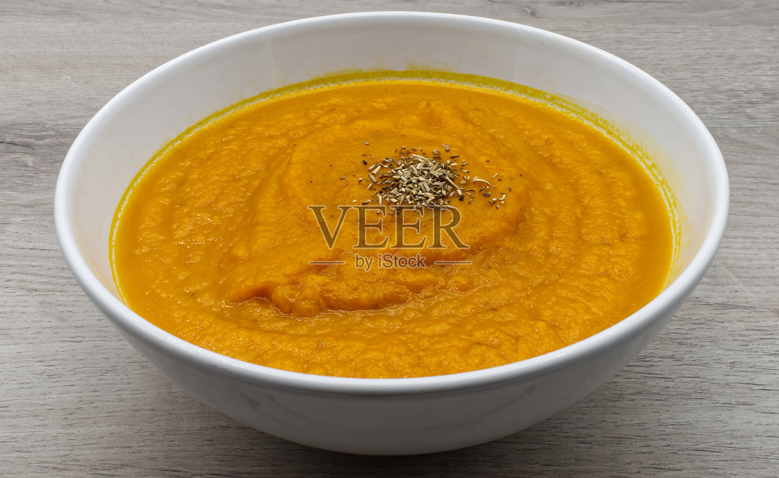 一碗美味的浓郁橙色奶油汤，可能是用南瓜或胡桃南瓜制作的，精美地装饰着芳香的干香草，摆放在浅色木质表面上。照片摄影图片