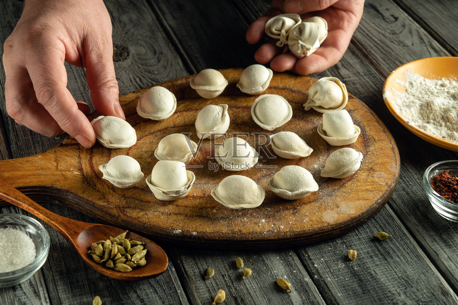 熟练的双手在温暖的厨房里将新鲜制作的饺子整齐地摆放在木板上。旁边，各种香料和面粉增添了烹饪的氛围。照片摄影图片