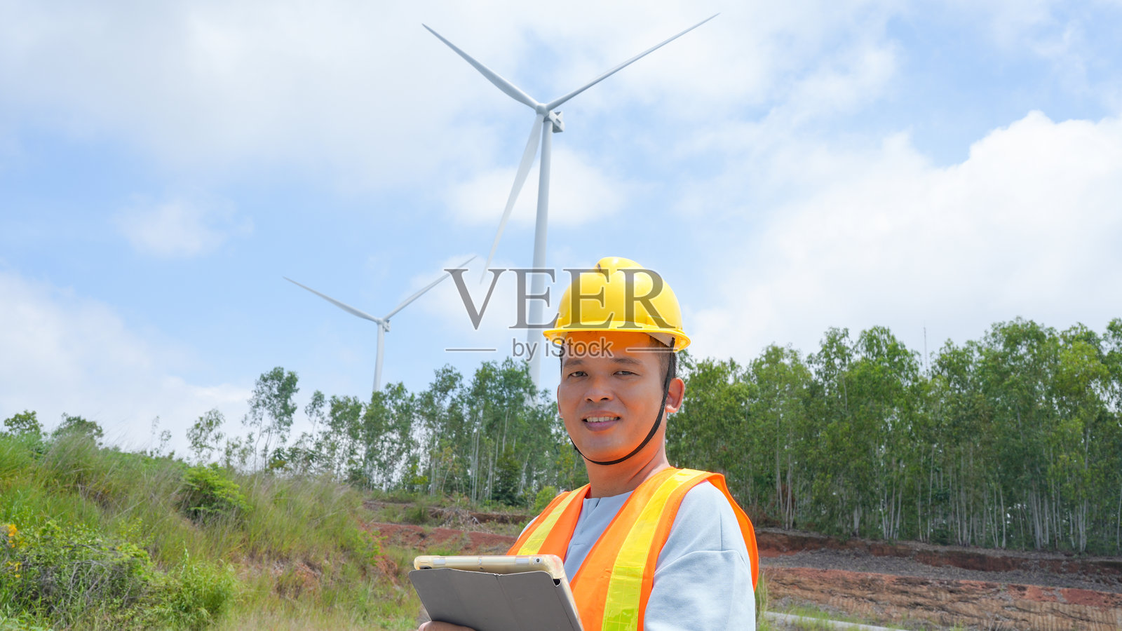 微笑的工人在安全头盔和马甲的保护下，站在风力涡轮机前，手持平板电脑。场景传达出可再生能源中的进步与可持续性的感觉。照片摄影图片