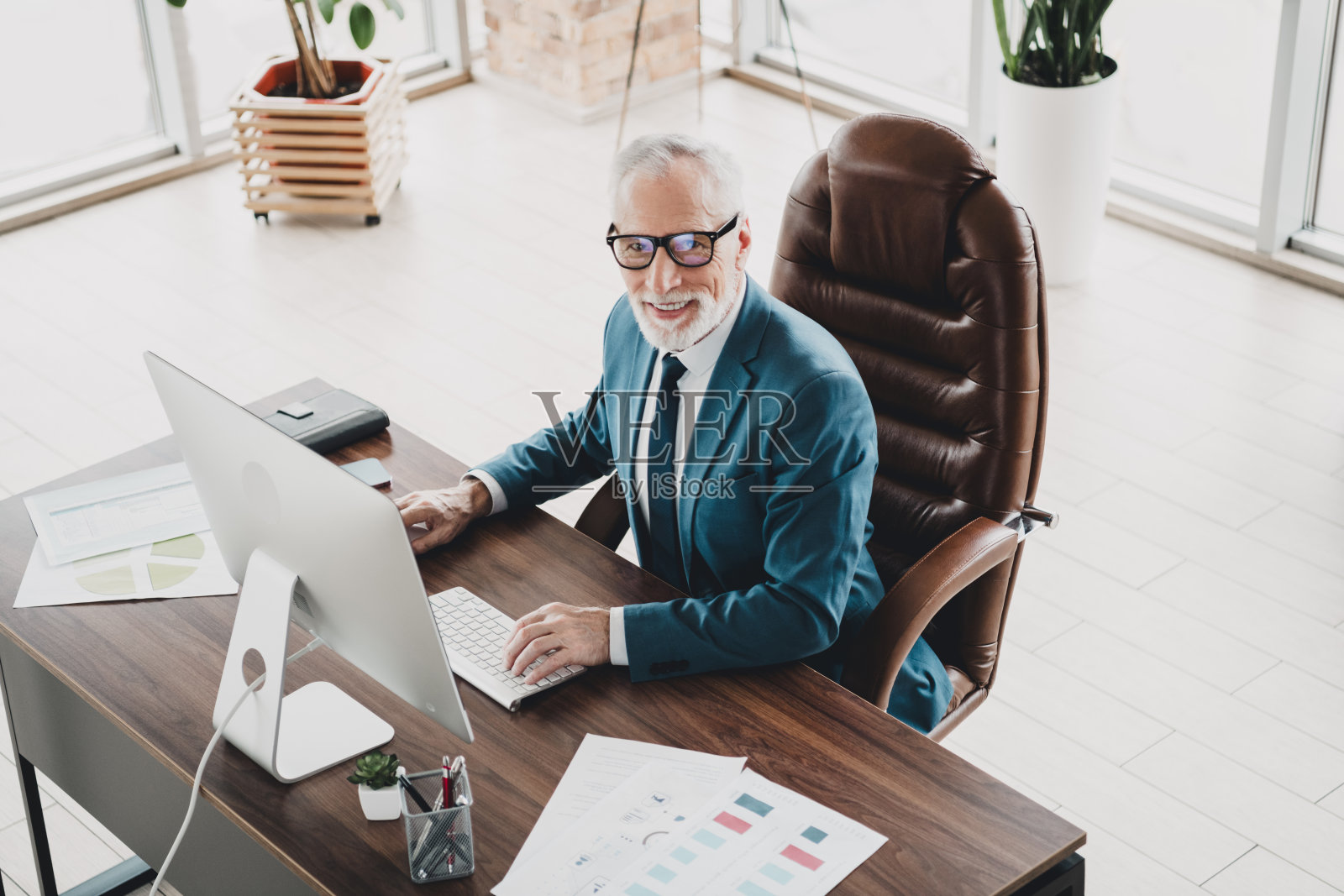 成熟的商人在现代办公室的桌子前自信地工作，身穿正式西装，旁边有电脑和文件。照片摄影图片