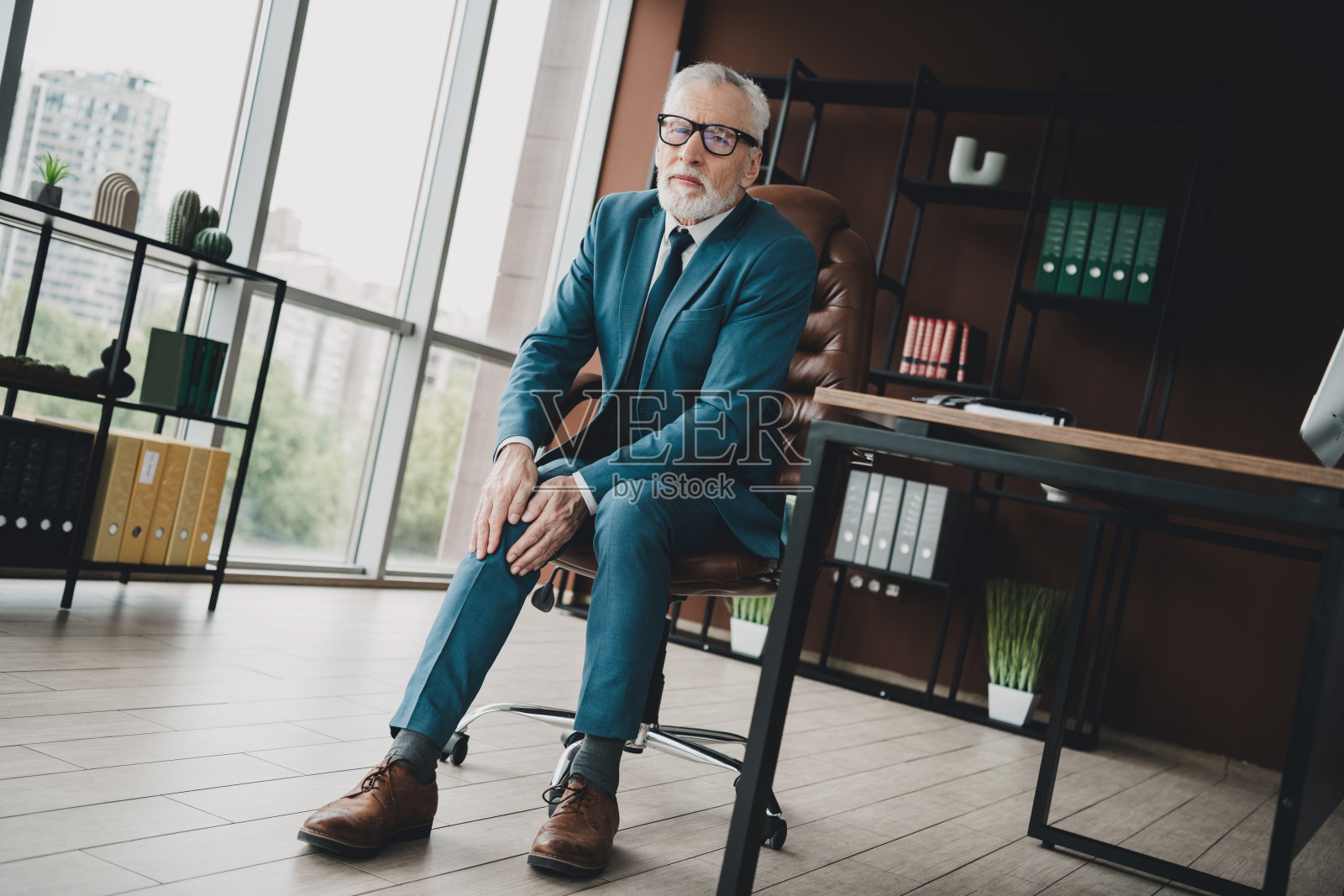 成熟的商人在时尚的正装中自信地坐在现代办公室的工作空间里，周围有装饰和大窗户。照片摄影图片