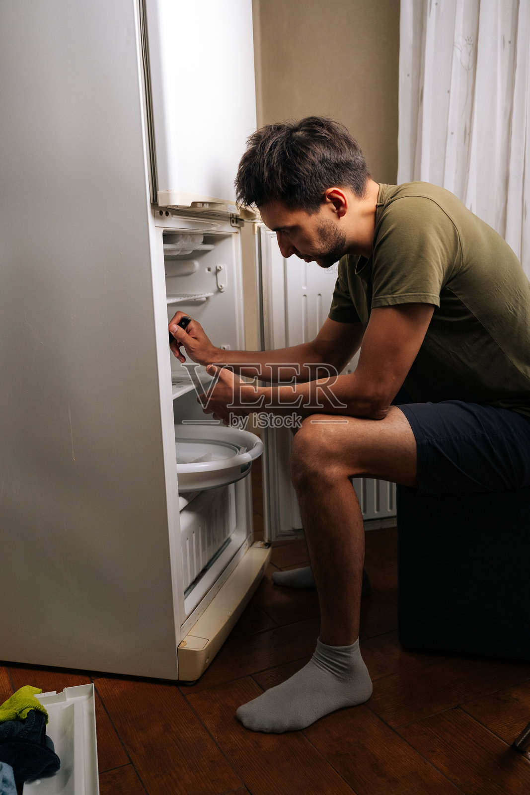 专注的年轻男子坐在凳子上，正在解冻冰箱并清洁内部，进行厨房的必要维护。照片摄影图片