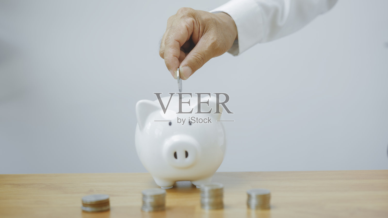 一个男人的手正在把硬币放入一个白色的储蓄罐。面前是一排硬币。储蓄退休金和存入基金的概念。金融与投资。照片摄影图片