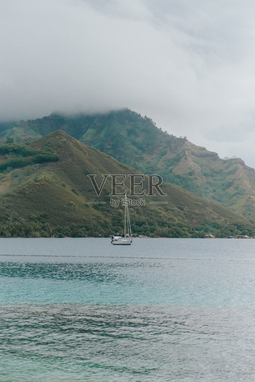 停泊在宁静的碧绿色泻湖中的帆船，背景是摩雷阿岛（法属波利尼西亚）郁郁葱葱的绿色山脉和戏剧性的云彩。照片摄影图片