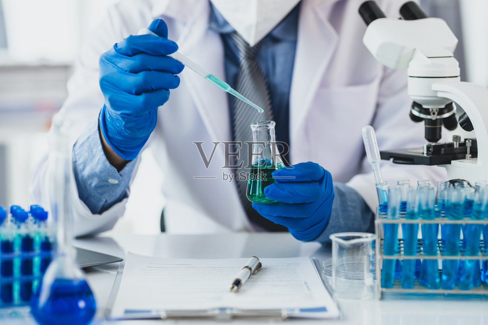科学家在实验室环境中分析液体。科学家手持一个装有蓝色液体的锥形瓶，在配备显微镜和试管的实验室中进行研究。照片摄影图片