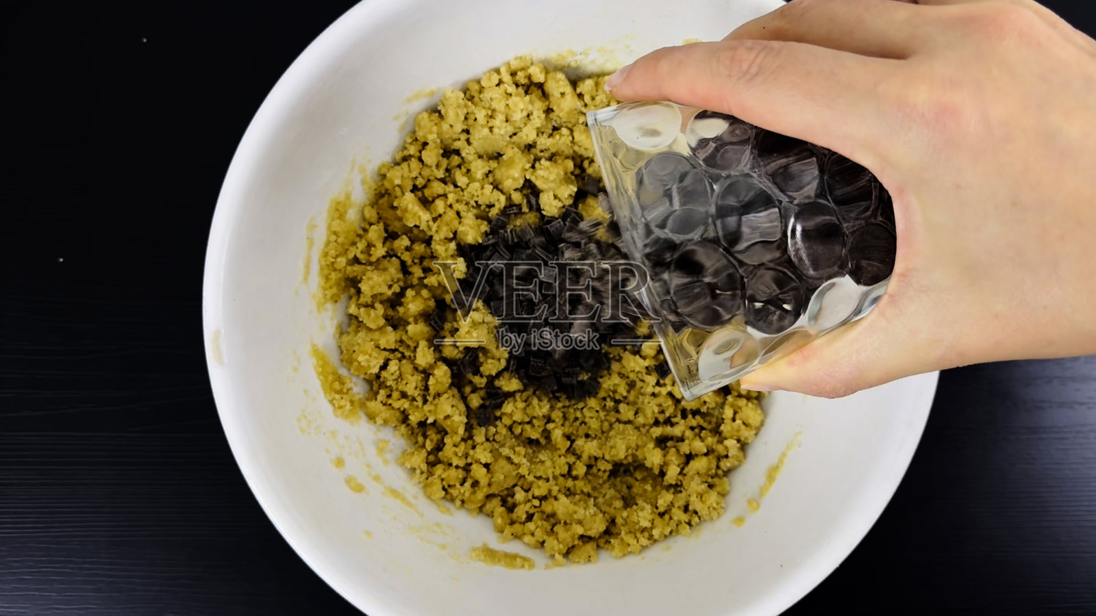 在黑色桌子上，手从玻璃杯中将巧克力豆倒入白色碗中的饼干面团，正在制作自制巧克力豆饼干的过程中。照片摄影图片