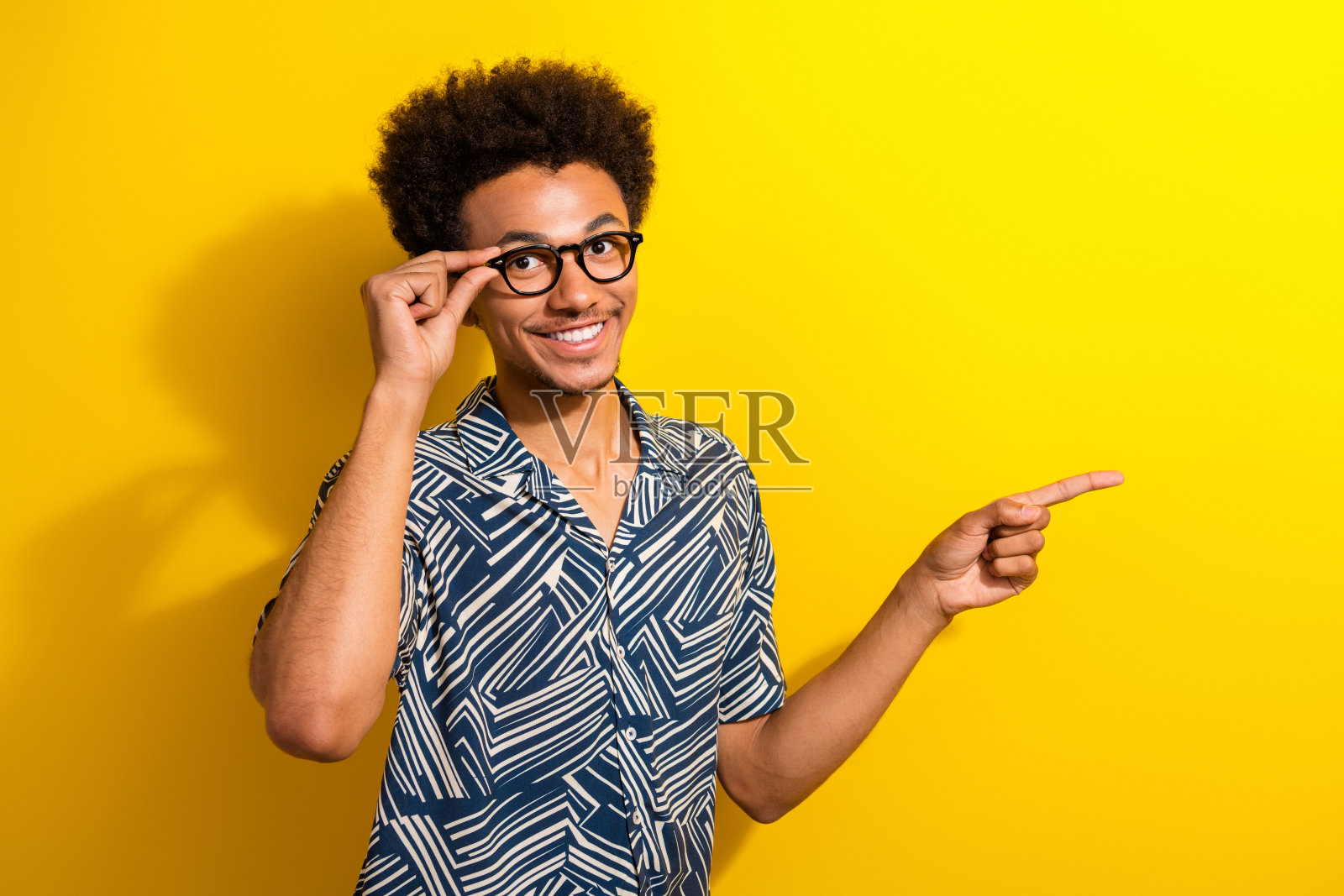 戴眼镜的快乐年轻男子在黄色背景下用手指着，展示一件时尚印花衬衫。照片摄影图片