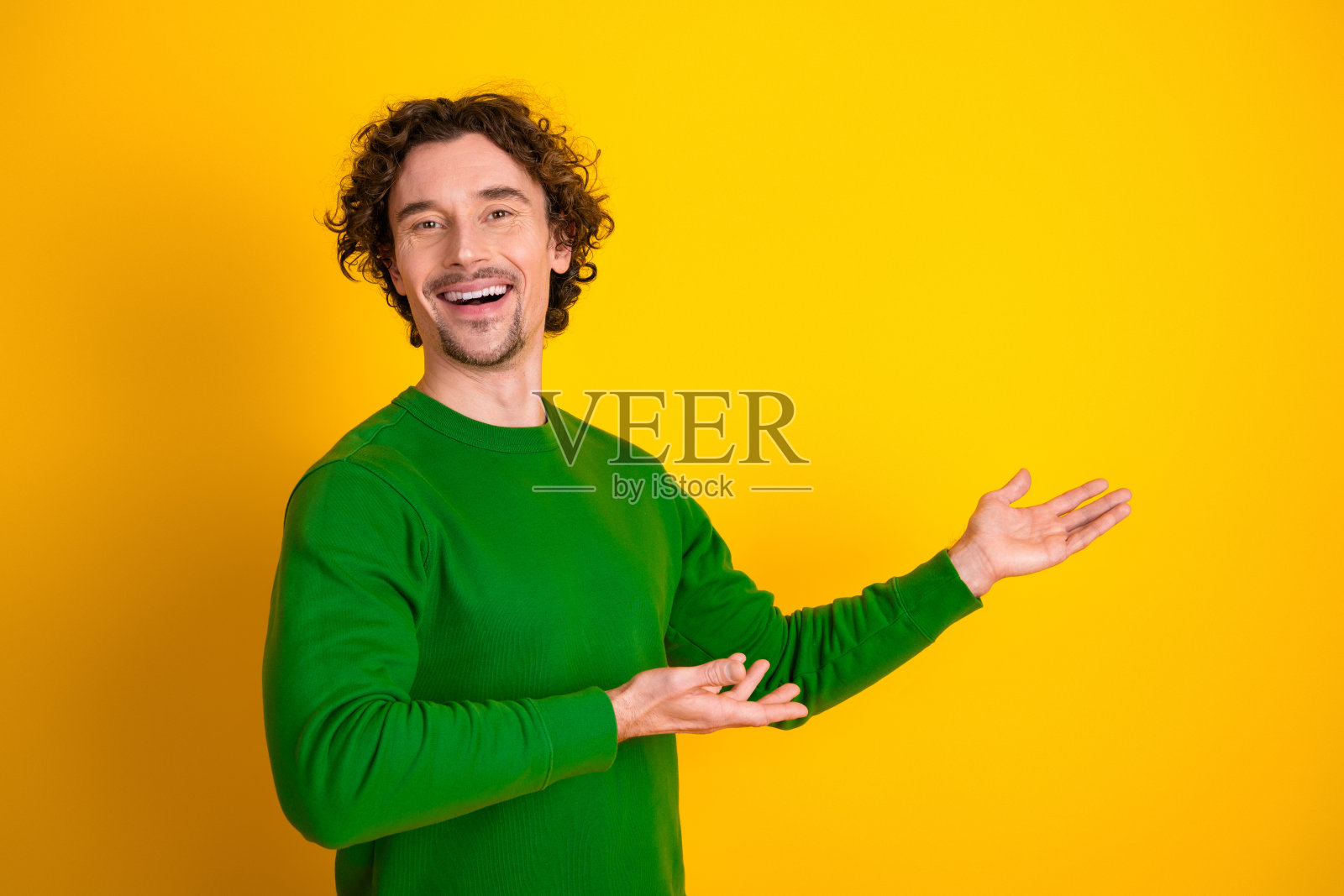 穿着绿色毛衣的年轻男子，卷发自信地在黄色背景前做手势，面带愉快的表情。照片摄影图片