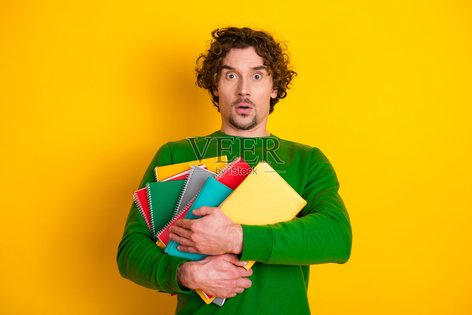 穿着绿色毛衣的年轻男子在黄色背景前手持五彩缤纷的笔记本，惊讶地睁大眼睛，张嘴表达惊讶。照片摄影图片