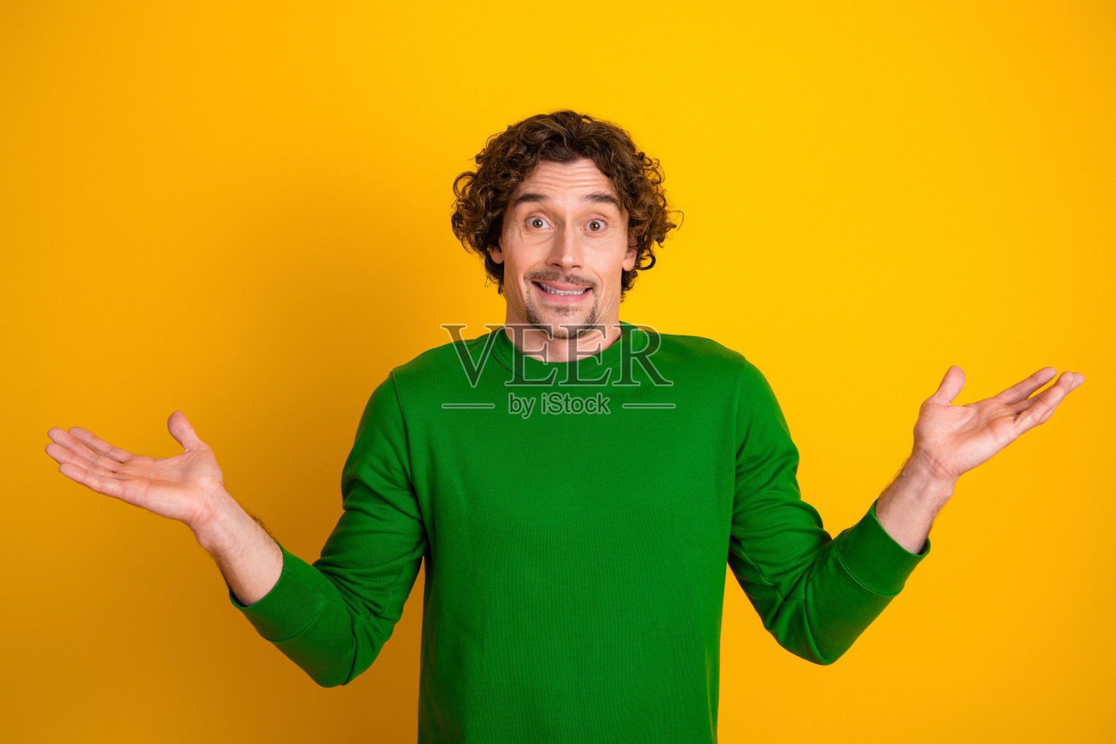 年轻男子穿着绿色毛衣，在黄色背景前做出不确定的手势，表达无忧无虑的心情和休闲风格照片摄影图片