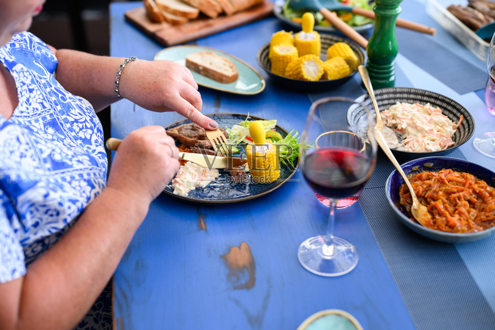 一位无法辨认的女性正在午餐盘上切割烤肉，盘中还有玉米、沙拉、红酒和切片面包，色彩丰富的烧烤餐点布置。照片摄影图片