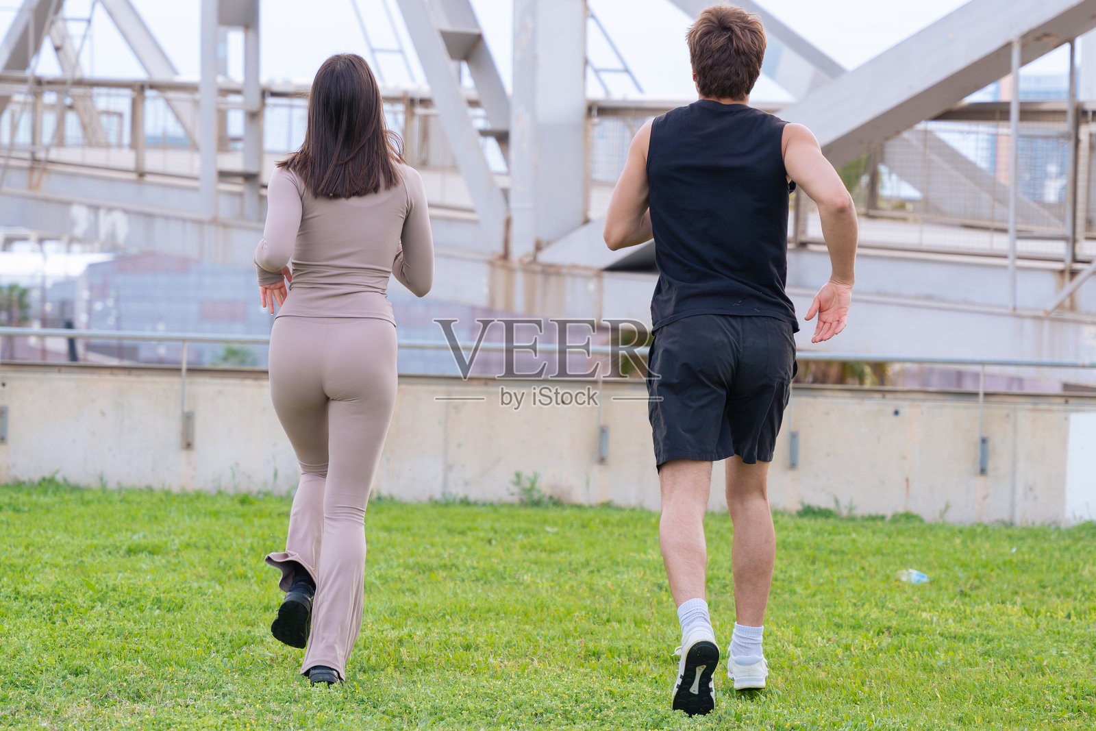 年轻的男女在大桥下的绿草地上一起慢跑照片摄影图片