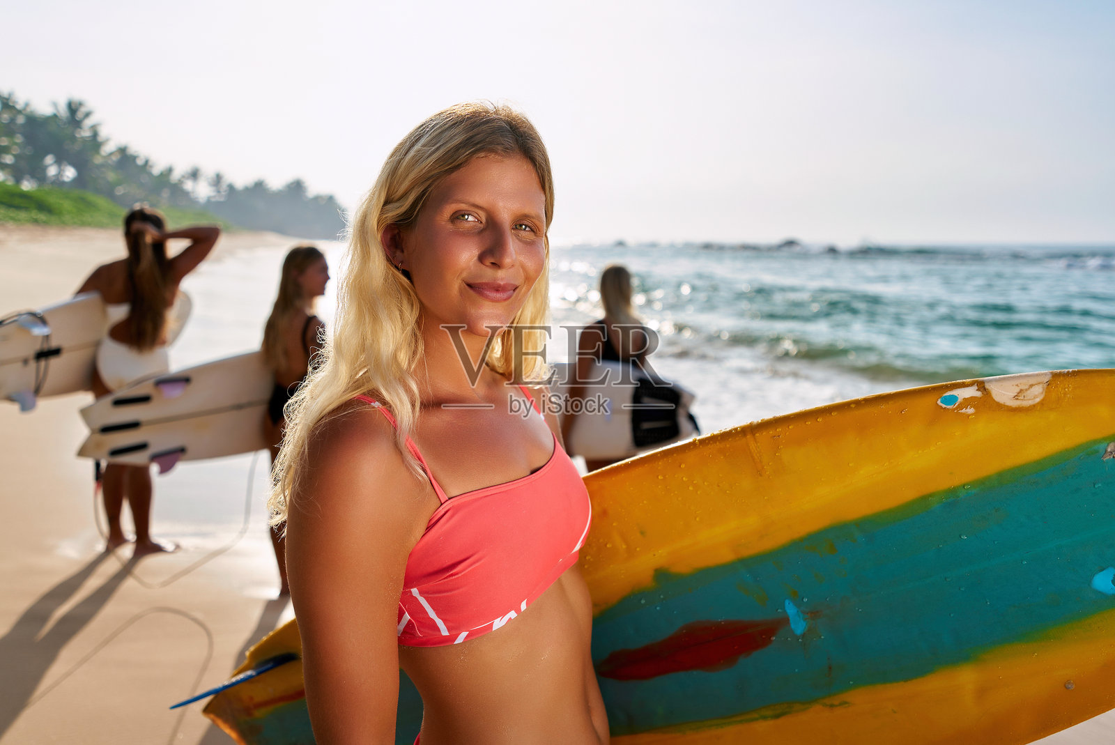 金发女性手持冲浪板在沙滩上微笑。一群冲浪者在海浪附近学习冲浪。中年和老年人进行冲浪训练。适合各个年龄段的冲浪学校。阳光明媚的海边日子。照片摄影图片