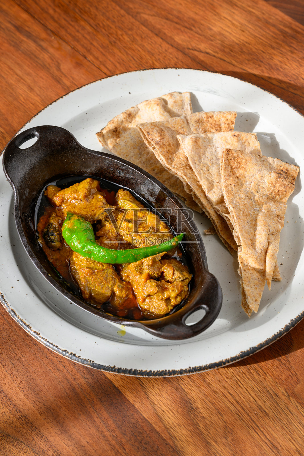 黑色铸铁锅中的羊肉咖喱，旁边配有全麦烤饼，放在古董胡桃木桌上。照片摄影图片