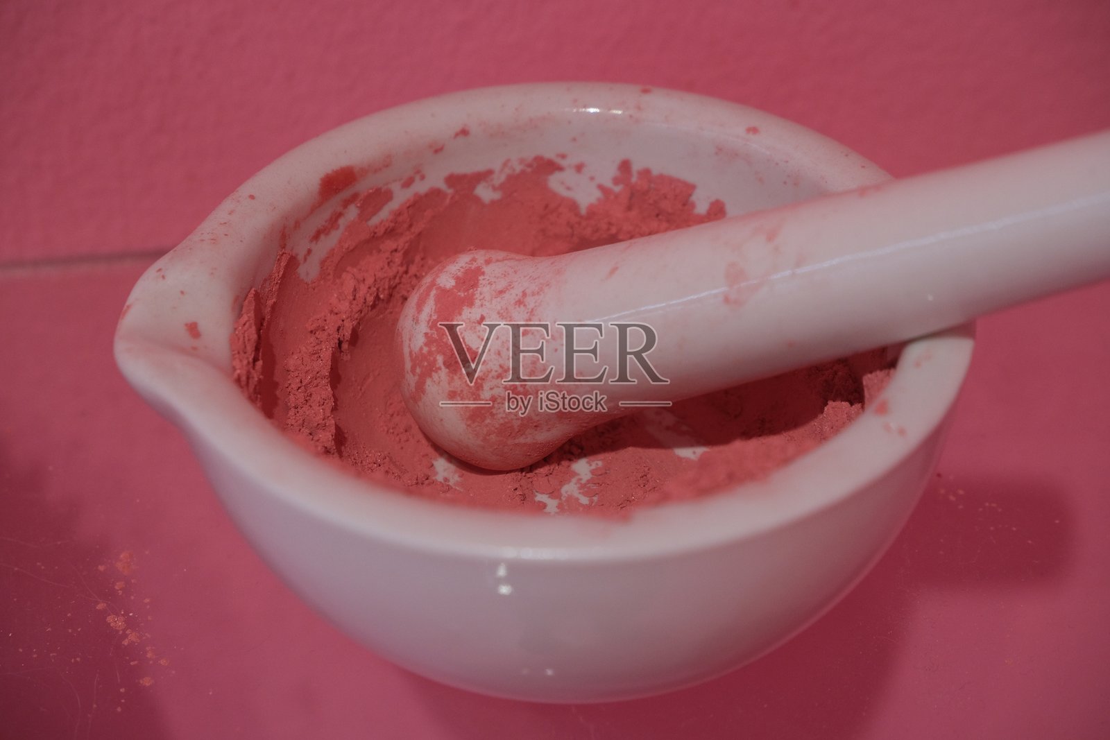 一个白色陶瓷研钵和研杵装满粉红色粉末，放在一个匹配的粉红色表面上，暗示着化妆品或颜料。照片摄影图片