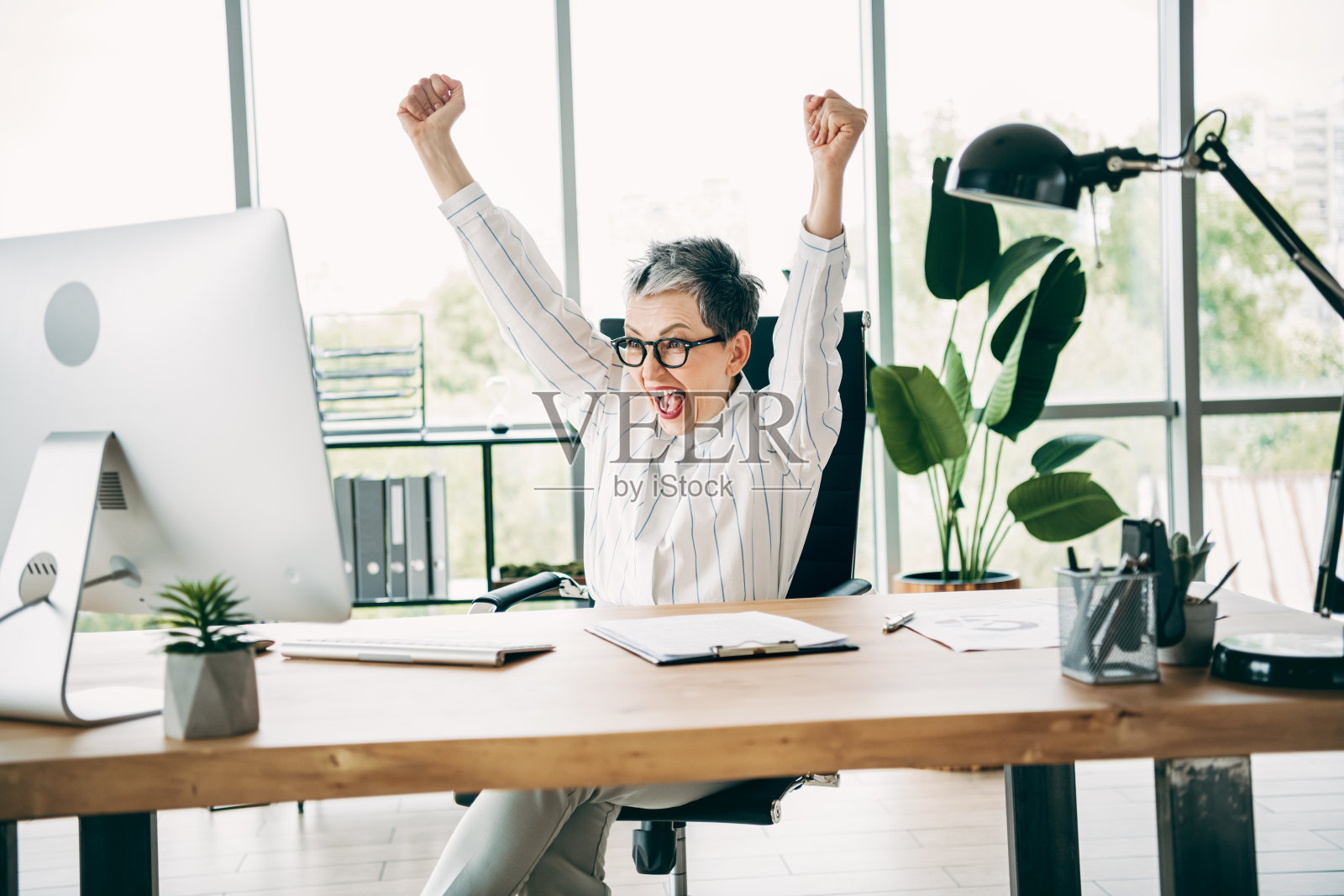 自信的女商人在现代办公室中庆祝成功，周围有电脑和文件，室内设计时尚。照片摄影图片