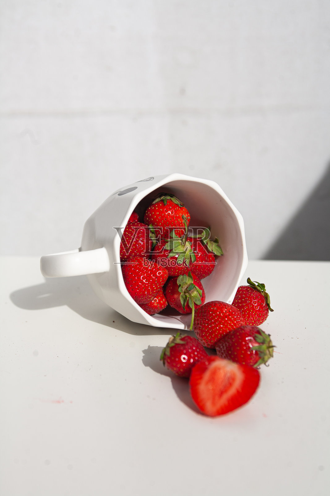 白色表面上的碗和洒落的成熟草莓杯。光滑的红色浆果和绿色顶部在正午的光线下投射出清晰的阴影，唤起夏日的清新、简单的丰盈和干净的极简美学。照片摄影图片