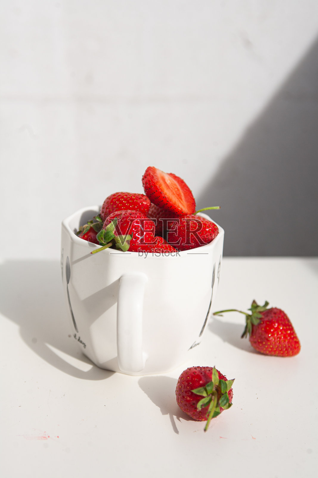 白色表面上的碗和洒落的成熟草莓杯。光滑的红色浆果和绿色顶部在正午的光线下投射出清晰的阴影，唤起夏天的清新、简单的丰盈和干净、极简的美感。照片摄影图片