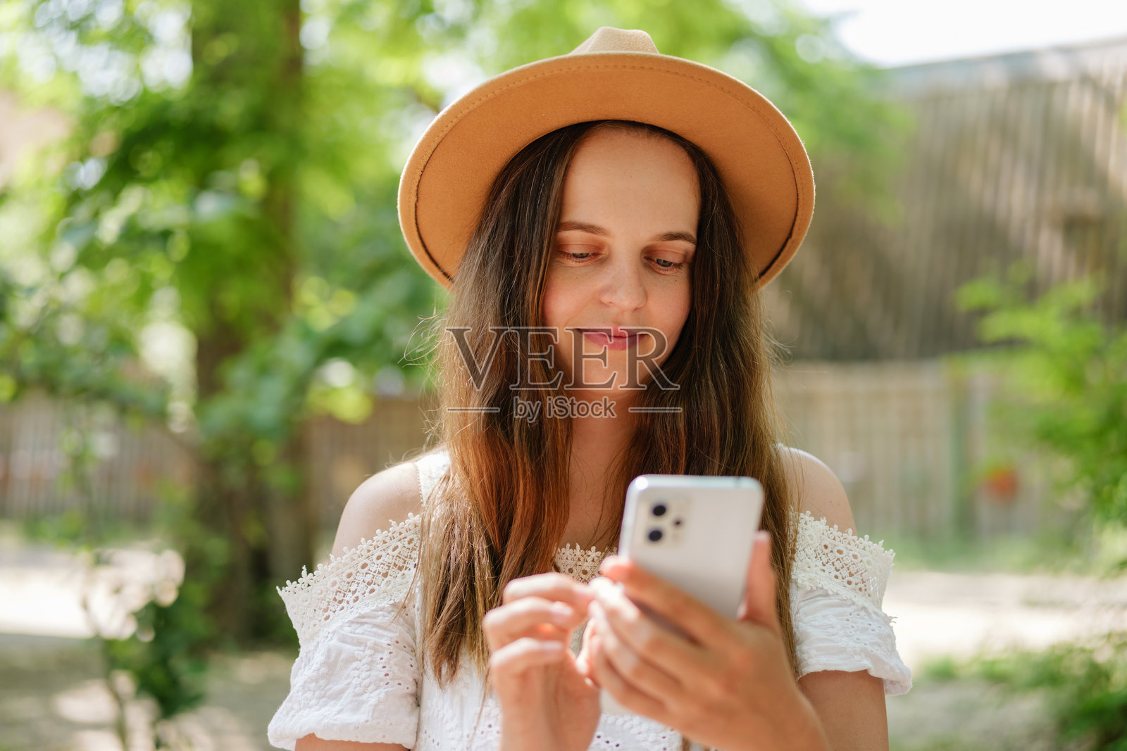 一位年轻女性穿着白色露肩上衣和草帽，微笑着低头看着她的智能手机，身处绿叶环绕的户外。照片摄影图片