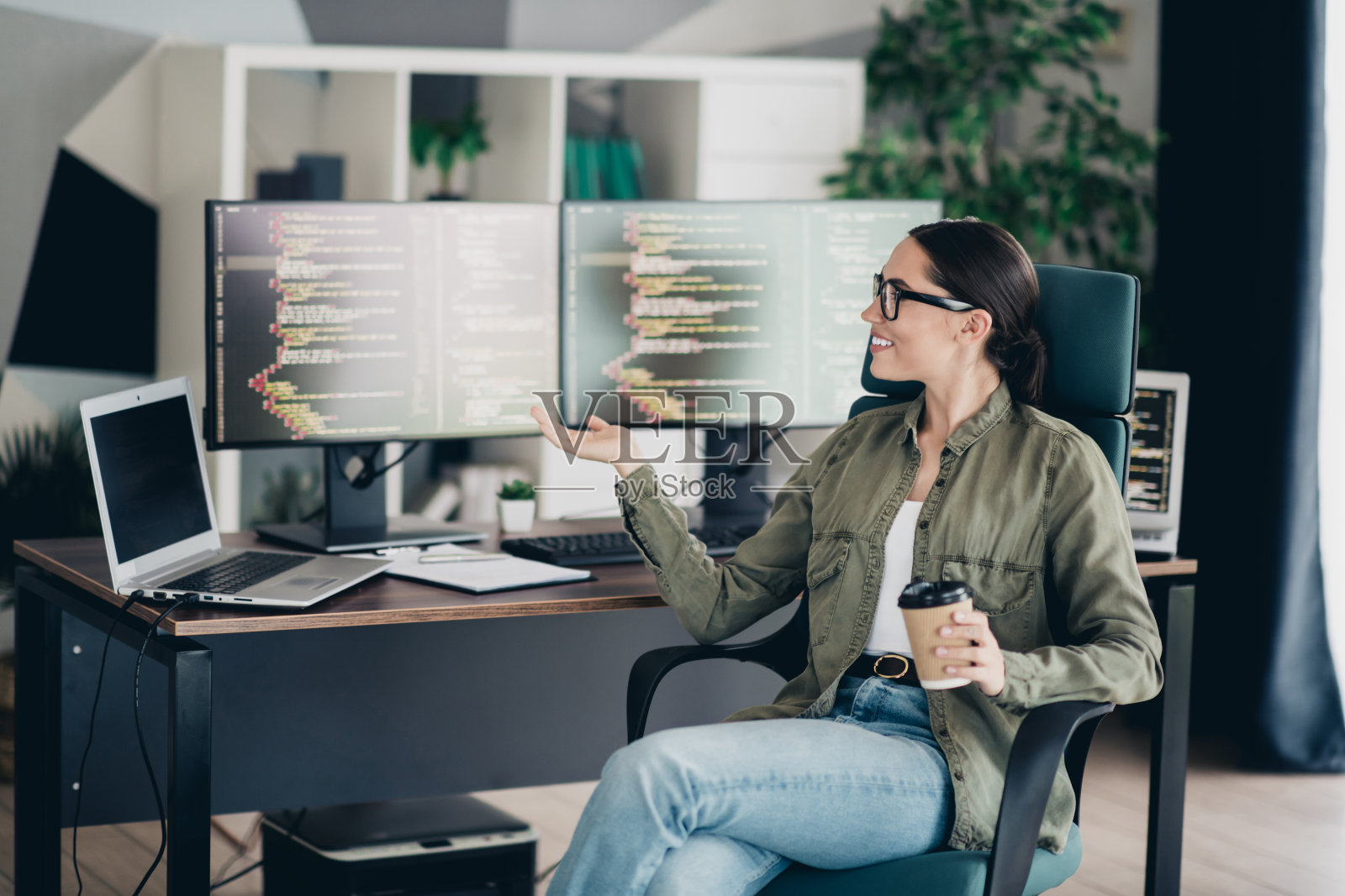 年轻的女性程序员在舒适的工作空间里，周围有多台显示器和笔记本电脑，手里拿着一杯咖啡。照片摄影图片
