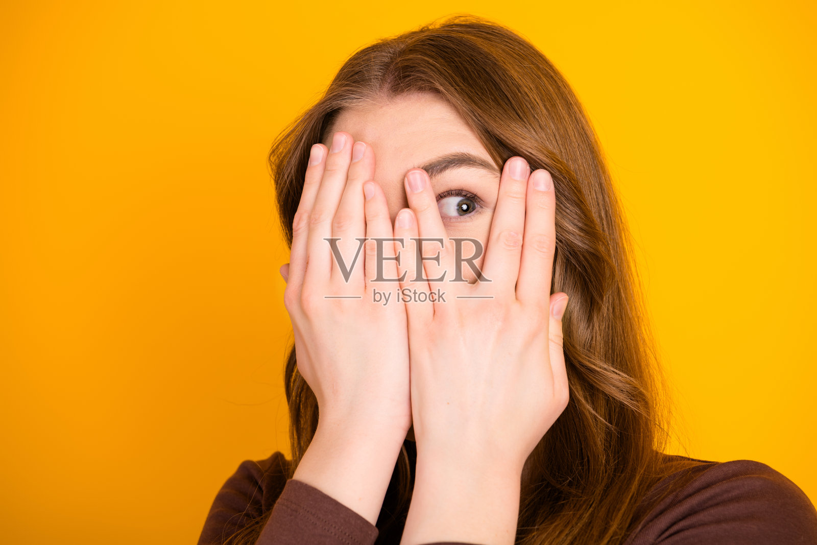 一位露出一只眼睛的女性透过双手窥视，背景为鲜艳的黄色。照片摄影图片
