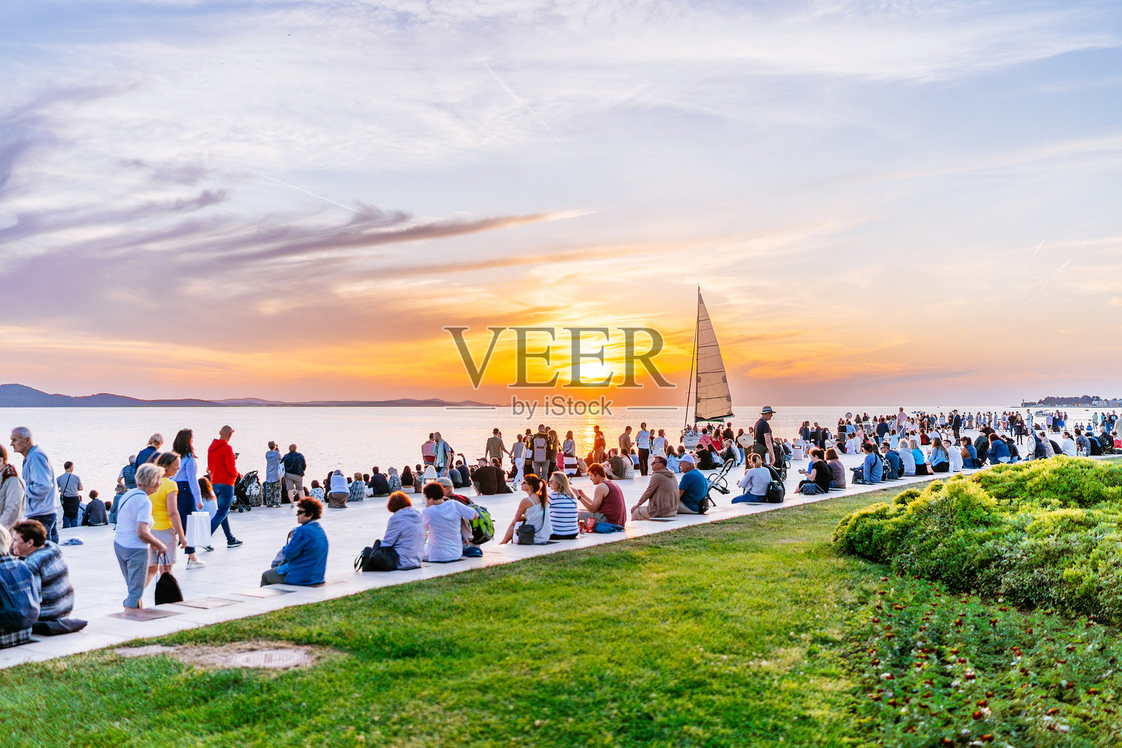 人群坐在扎达尔海滨长廊上，观看亚得里亚海的日落和帆船。照片摄影图片