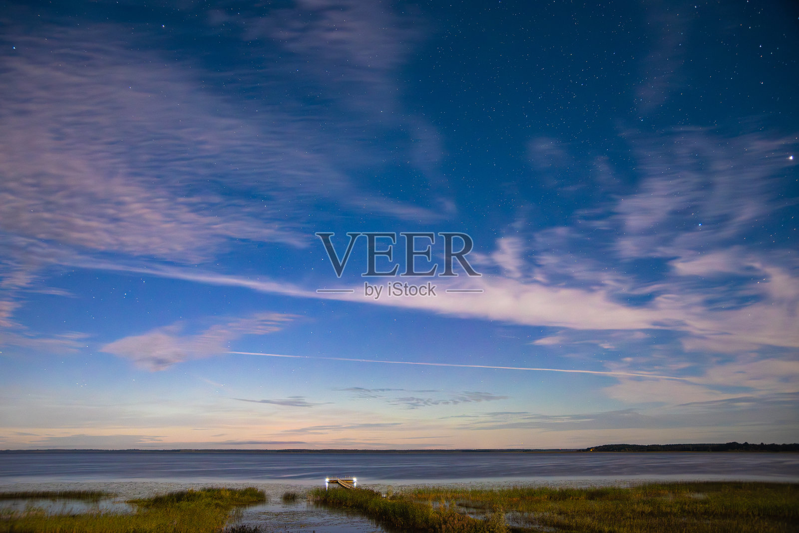 宁静海面上的夜空，散布着云朵、星星，以及小码头上方微弱的光迹。照片摄影图片