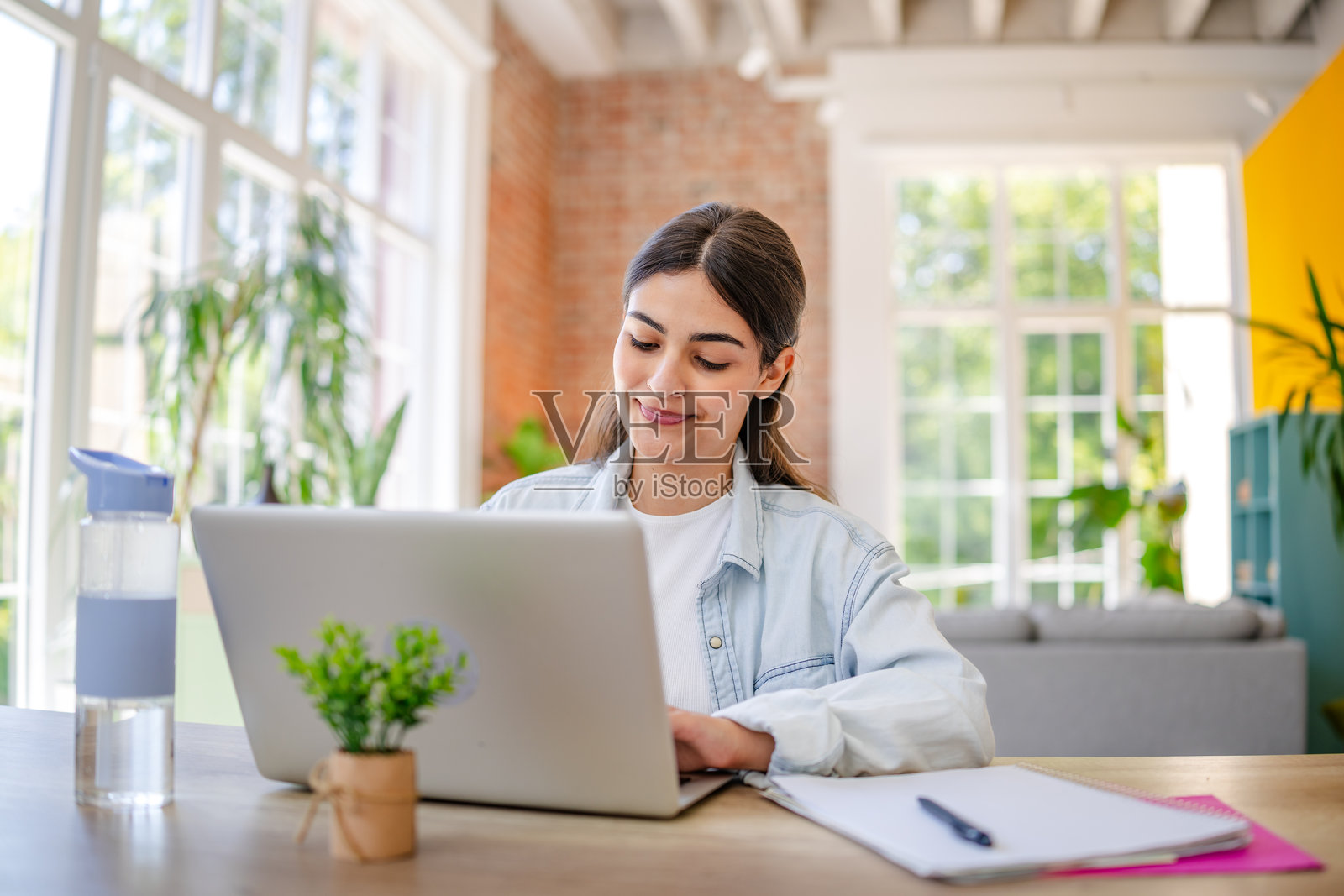 年轻女性在明亮的客厅中享受高效的居家办公时光，周围布满植物和自然光。照片摄影图片
