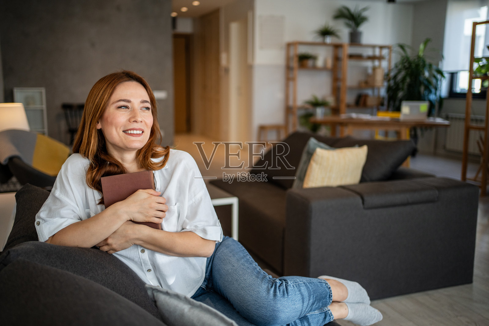 年轻女性在沙发上放松，手里拿着一本书，沉浸在白日梦中照片摄影图片