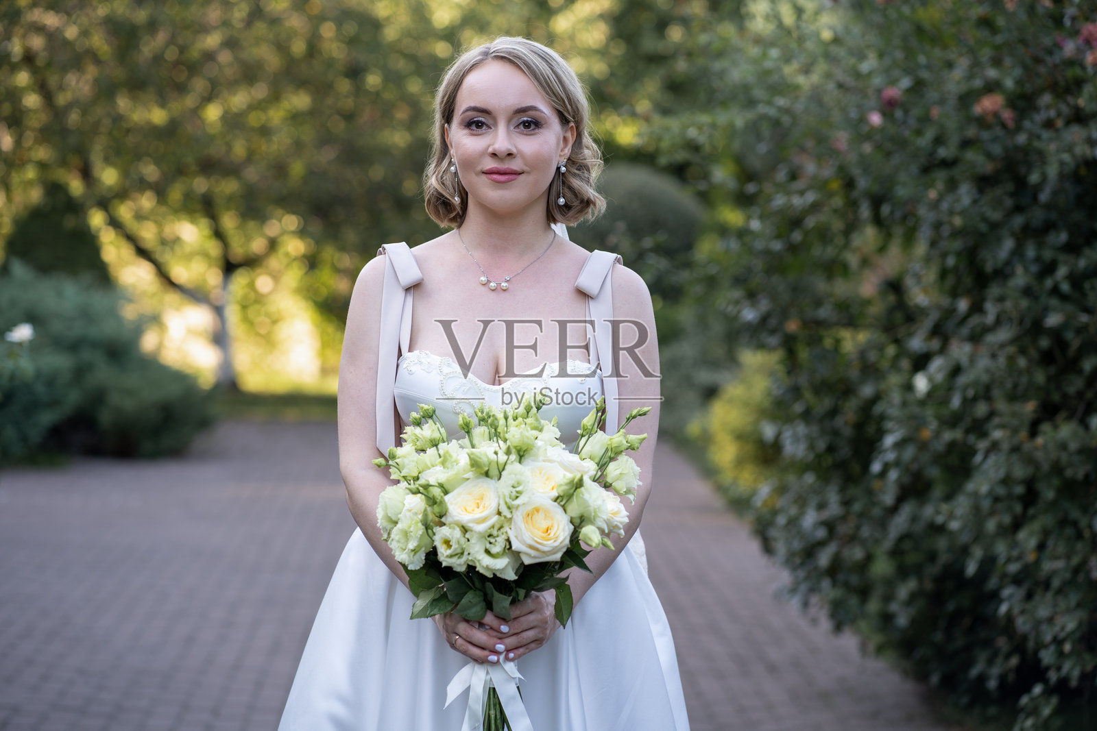 身穿白色茶长婚纱的新娘正面肖像，轻柔地捧着一束象牙色和淡粉色玫瑰及绿色花蕾。拍摄于户外的鹅卵石小径上，四周环绕着绿意，散发出永恒的优雅和宁静的婚礼美感。照片摄影图片