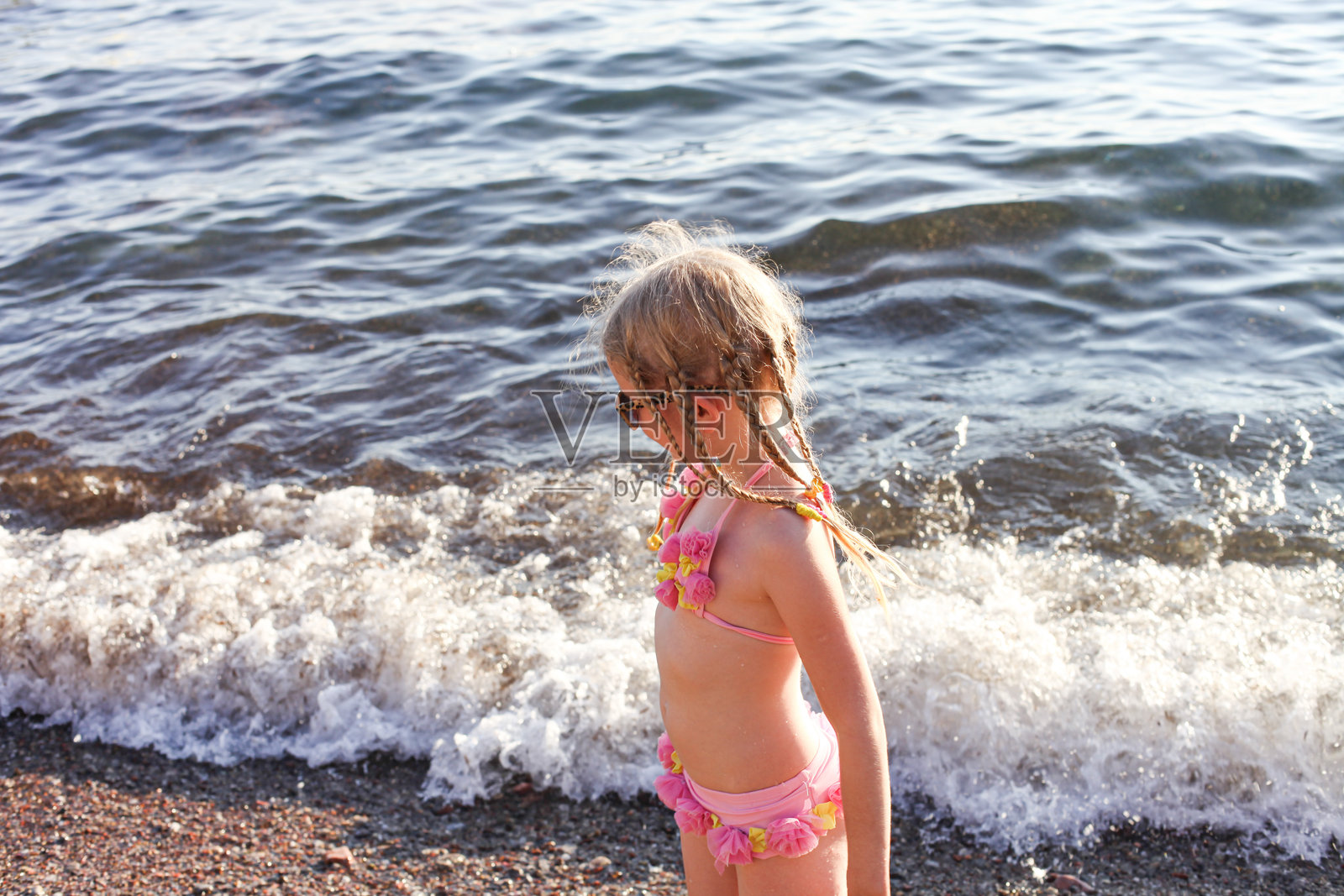 侧面视图的7岁白人女孩，扎着辫子，望着海浪。孩子穿着粉色泳衣，戴着太阳镜，站在沙滩上。照片摄影图片