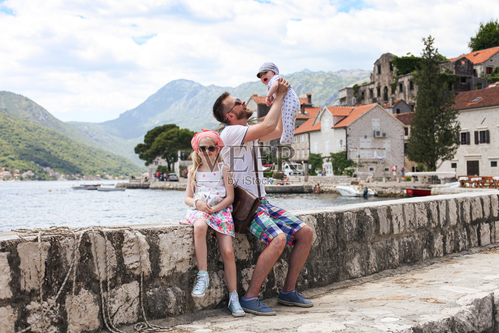 年轻父亲与两个孩子坐在黑山的佩拉斯特水边。男人与婴儿儿子和女儿被海洋、山脉和历史建筑环绕。照片摄影图片